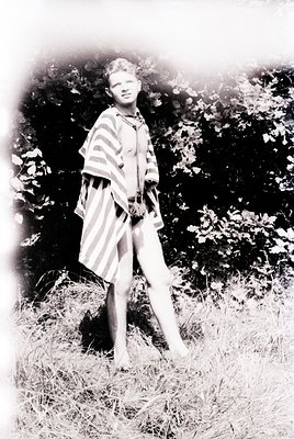 Vintage black-and-white portrait of a young person in a striped dress and suspenders, standing outdoors among tall grass and ...