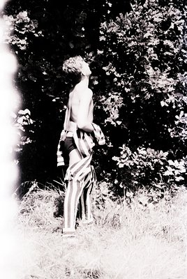 Vintage black-and-white photo of a shirtless child in striped pants, standing outdoors among tall grass and dense foliage. Th...