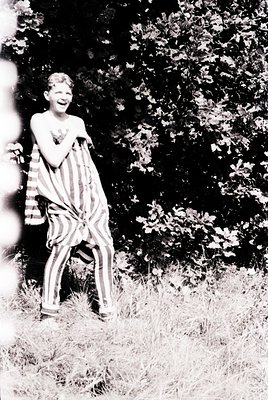 Mid-20th century black-and-white photo of a young boy in a striped prison-style outfit, posing joyfully outdoors. The setting...