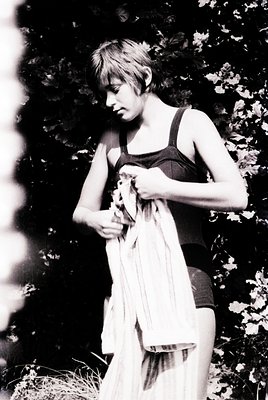 Vintage black-and-white portrait of a woman in a loose, draped swimsuit holding a towel, standing outdoors among leafy greene...