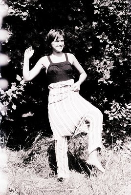 Young woman in 1970s-style cropped top and flared pants poses outdoors among dense foliage. Her relaxed stance and bright smi...