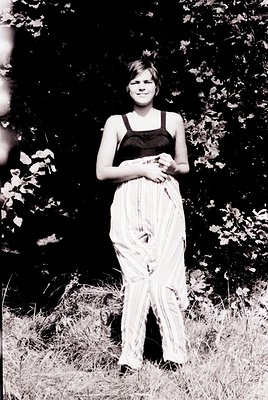 Portrait of a woman in a 1970s-style dress—black top with sheer, layered white skirt—standing outdoors among dense foliage. N...