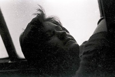 Black-and-white portrait of a person resting their head on a window ledge, framed by the interior of what appears to be a tra...