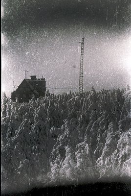 A vintage black-and-white photo of a remote alpine cabin nestled among rugged, snow-dusted terrain. The single-story structur...