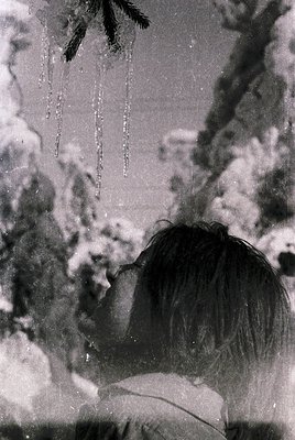 Black-and-white winter scene: child’s back facing camera, bundled in heavy coat, hands near mouth under falling snowflakes. D...