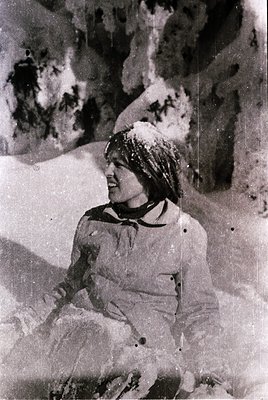 Vintage black-and-white photo of a person skiing in alpine terrain, mid-laugh, wearing a knit cap and ski parka. Snow-covered...