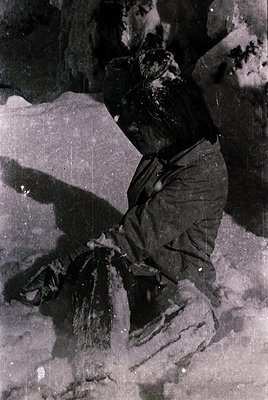 Indoor scene featuring a person seated on a stone floor, holding a bundle wrapped in dark fabric. The setting appears rugged,...