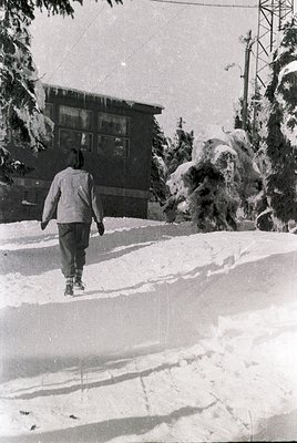 Mid-century winter scene featuring a lone figure in a light jacket and trousers trudging through snow toward a two-story buil...
