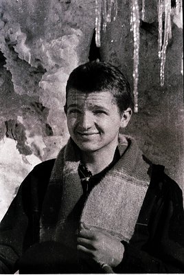 Portrait of a young man in a checkered scarf and dark jacket, posed against a textured, possibly cave or rock wall with icicl...