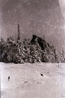 Vintage black-and-white alpine village blanketed in snow, with a prominent wooden church and communication tower. Snow-covere...