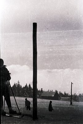 Vintage black-and-white photo of a photographer using a large tripod camera in a rural setting, likely mid-20th century. The ...