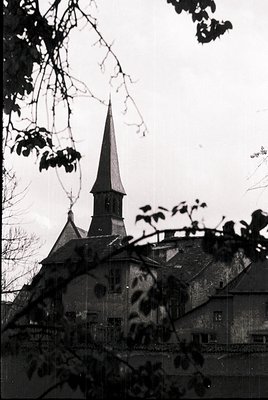 Mid-20th century church with steep gable roof and pointed steeple, framed by bare branches. Brick facade shows weathering, su...