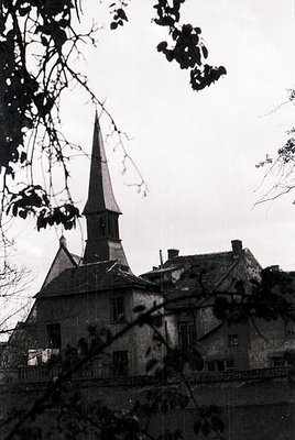 Historic stone church with steep spire and slate roof, framed by bare winter branches. Architectural details include small re...