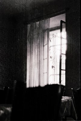 Vintage black-and-white interior shot featuring a partially open window with sheer curtains and a wooden frame. Sunlight stre...