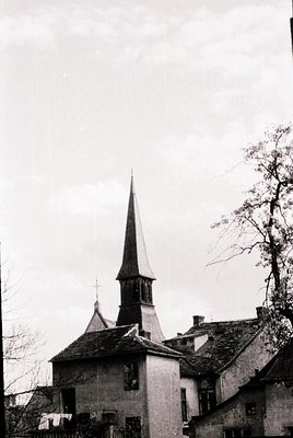 Historic church steeple with pointed spire and slate roof, flanked by weathered stone buildings. Likely European rural archit...