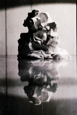 Double-exposure portrait of a person holding a hat, reflected in a mirror or glass surface. The composition creates a ghostly...