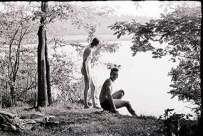 Black-and-white candid of two men by a serene lakeside, mid-20th century. One kneels in swim trunks, the other stands shirtle...