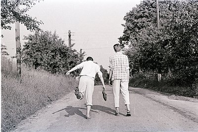 Two men in mid-stride on a rural road, 1960s-era clothing (short-sleeve button-up, khakis, loafers). Lush greenery and utilit...