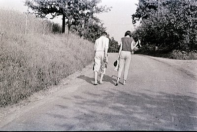 Two individuals walk away from the camera on a rural road, one holding a ball. Mid-20th century clothing suggests or . Lush g...