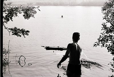 Black-and-white shot of a man standing in shallow water, facing a distant swimmer in a lake or river. Overgrown foliage frame...