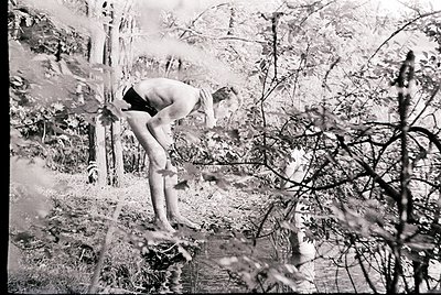 Black-and-white shot of a shirtless man mid-dive into a shallow, wooded pond, framed by bare branches. Dynamic composition ca...