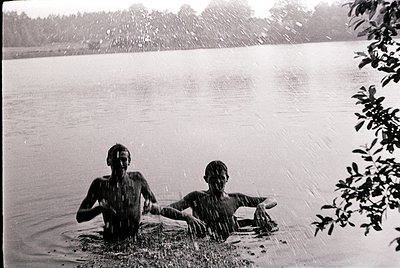 Two shirtless boys wade through shallow water in a mid-20th-century lake setting, rain falling around them. Lush greenery fra...