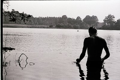 Black-and-white shot of a lone male wading in shallow water, back to camera. Dense forest line in background suggests a natur...