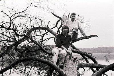 Black-and-white photo of three individuals perched on a fallen tree branch, likely mid-20th century. The man sits centrally i...