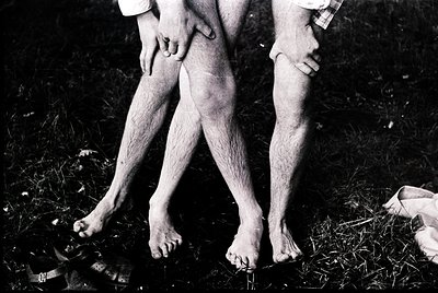 Black-and-white close-up of two barefoot legs intertwined, seated on grass. Clothing suggests 20th-century casual wear. Possi...