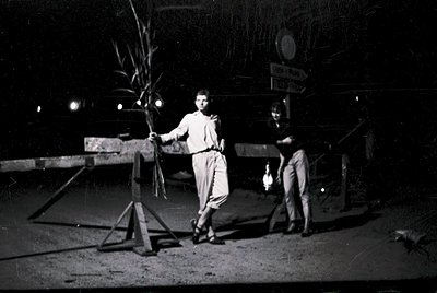 Black-and-white nighttime street scene featuring two individuals preparing a large palm frond for a festive display. The man ...