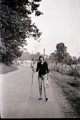 Mid-century woman in a rural setting, carrying a bundle of sticks and a cloth bag, walking along a paved path bordered by tre...