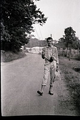 Young man in 1960s-era casual attire—plaid shirt, baggy trousers, and sandals—walks along a rural road. Holds a cane or walki...