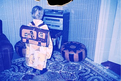 Vintage 1970s interior shot: child in patterned robe and backpack stands beside a large stereo console, facing a tiled wall w...