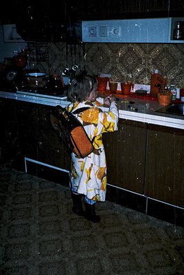 A young child in a patterned dress with a brown backpack stands at a mirrored kitchen counter, reaching for a red pot. Decora...