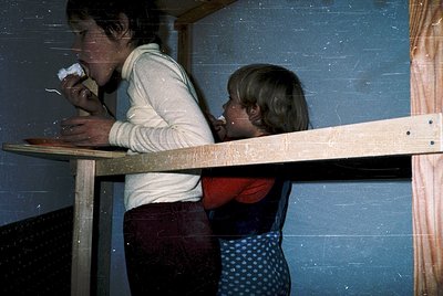 Two children in 1970s-era clothing share a meal at a wooden picnic table indoors, likely a family home. The boy wears a long-...