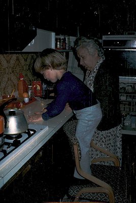 Vintage kitchen scene featuring two women preparing food, likely mid-1960s–70s. One woman kneels at a gas stove with a kettle...