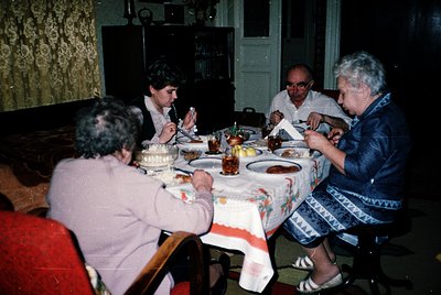 Family gathering at a round wooden table adorned with floral-patterned tablecloths, celebrating a birthday with cake and drin...