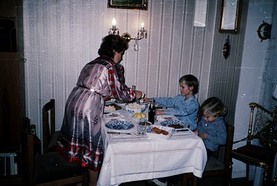 Vintage indoor family meal scene: woman in patterned dress serves two children at a table set with blue-and-white crockery, b...