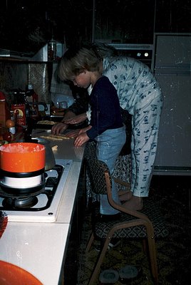 Mid-20th century kitchen scene featuring a woman in floral pajamas preparing food on a gas stove, 1960s-1970s. Orange kettle ...