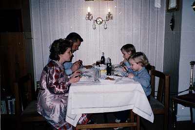 Family dinner scene in a mid-century interior, featuring three adults and two children seated around a wooden table. The tabl...