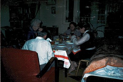 Vintage indoor gathering featuring five seated adults at a round table, adorned with patterned tablecloths and floral centerp...