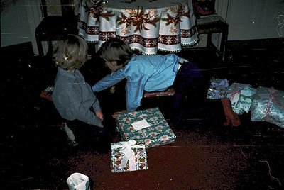 Vintage holiday scene featuring two children (early 1970s) opening wrapped gifts on a patterned rug. Decor includes floral-pa...