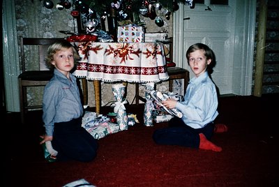 Two young boys kneel on a red carpeted floor, surrounded by wrapped gifts under a decorated Christmas tree. The boy on the le...