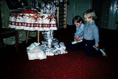 Two children in 1970s-style denim outfits kneel on a red carpeted floor, unwrapping wrapped gifts under a festive tablecloth ...