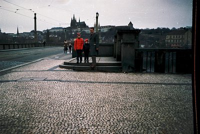 Vintage bridge scene with cobblestone pavement and overhead tram wires. Three figures—adults in winter coats and a child in a...