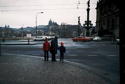 Three individuals stand on a cobblestone plaza near a historic fountain in a European city, likely Prague. The adult in a red...