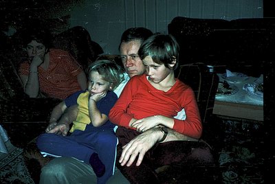 Vintage indoor family portrait from the 1970s, featuring three generations seated on patterned upholstery. Adults in long-sle...
