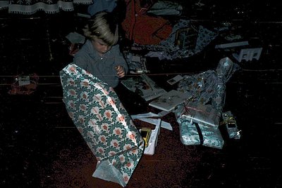 Child unwrapping holiday gifts in a dimly lit room, surrounded by wrapped presents and patterned wrapping paper. Mid-20th cen...