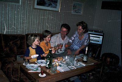 Family gathering in a dimly lit, rustic interior, likely Eastern Bloc-era (1970s–1980s). Four adults and two children seated ...