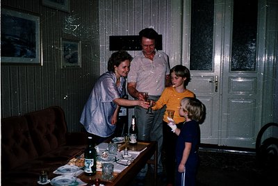 Family gathering in a dimly lit living room, likely mid-20th century. A woman in a blouse and apron serves drinks to two chil...